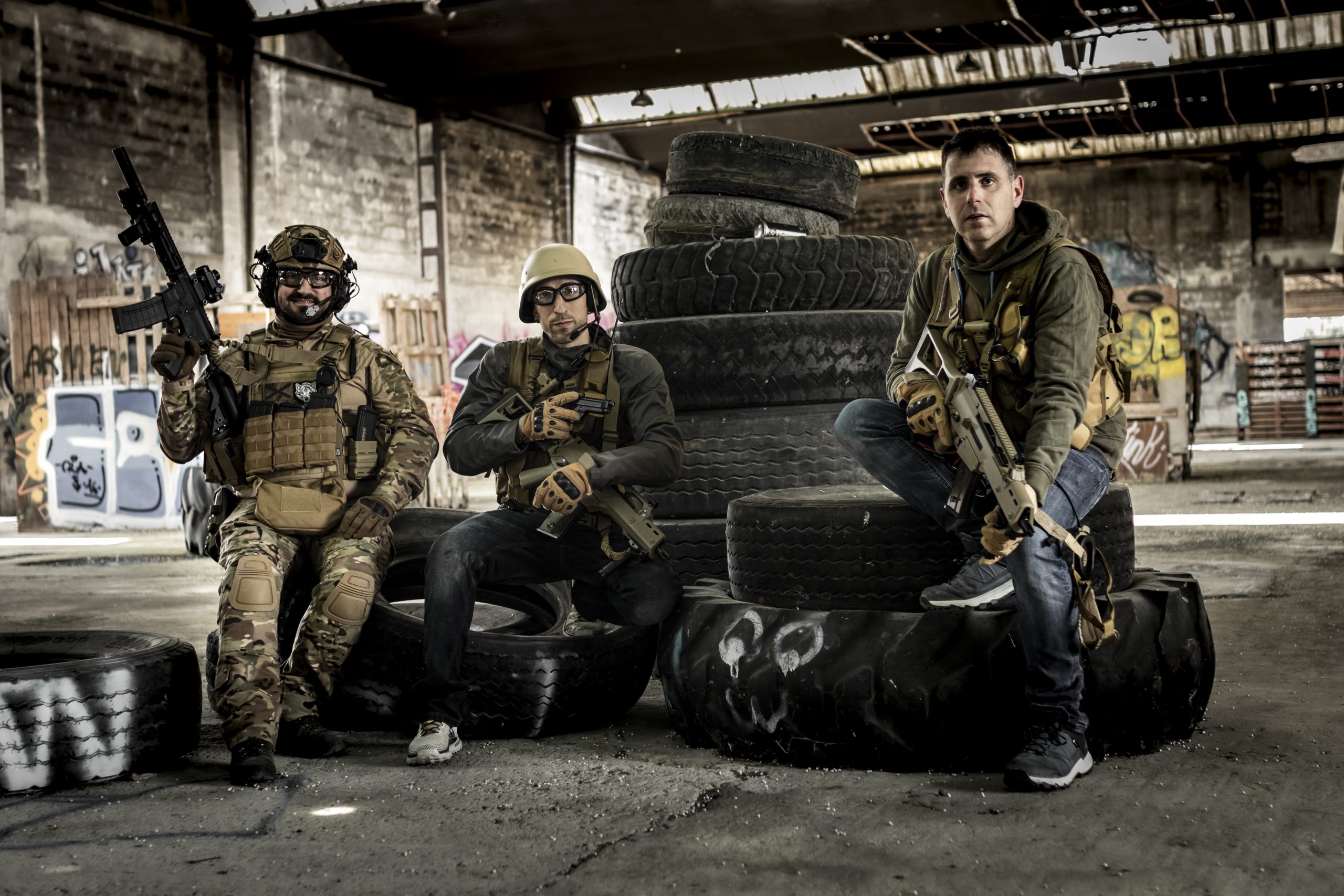 Tres jugadores de airsoft con equipación militar y réplicas en una nave industrial abandonada.