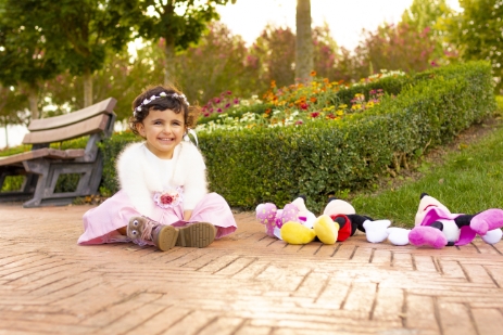 Niña pequeña sentada en el suelo de un parque, sonriente, con peluches a su lado.