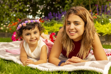 Madre y su hija pequeña tumbadas boca abajo sobre una manta en el césped de un parque, con flores y colorido al fondo.