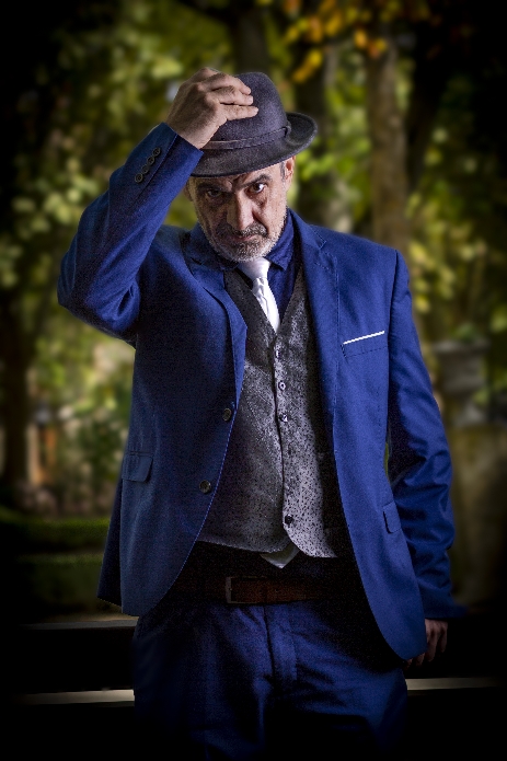 Oscar Balaperdida con traje azul y corbata blanca, quitándose el sombrero en un parque.