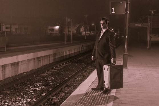 Retrato en tonos sepia de una persona con traje y maleta en el andén de una estación de tren.